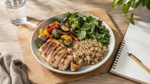 Balanced fat loss meal with grilled chicken, quinoa, roasted vegetables and fresh greens served on a light plate with a glass of water and notebook