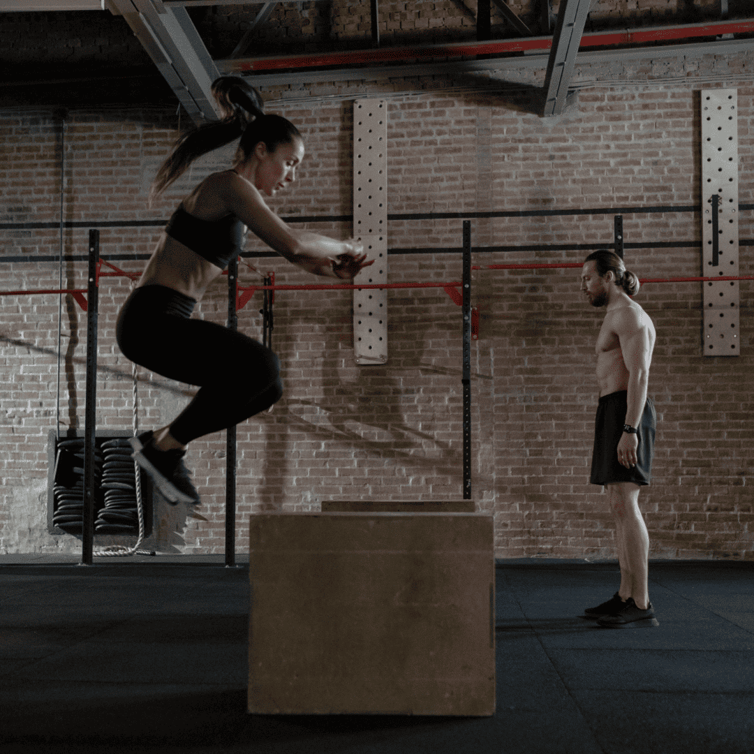 A man and woman workout in a crossfit box. The woman is completing box jumps while the man rests