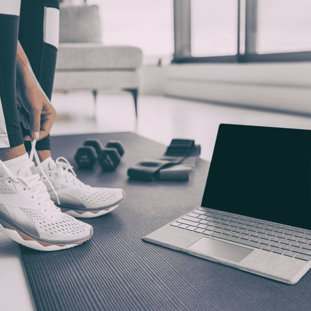 Close-up of a person tying their sneakers on a yoga mat, with a laptop and gym equipment like dumbbells and resistance bands nearby, representing the setup for an online workout or virtual training session.