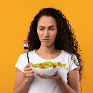 Curly-haired woman holding a bowl of salad, looking skeptical at a cherry tomato on her fork, against a vibrant orange background, depicting the struggle with appetite control and food choices