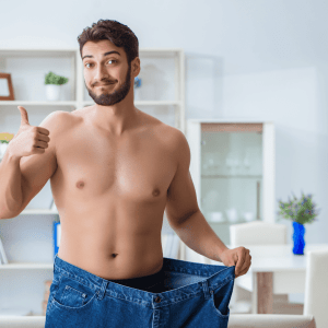 Smiling shirtless man giving a thumbs-up while holding out the waistband of his oversized jeans, symbolizing significant weight loss and personal achievement in a bright, modern living room setting.