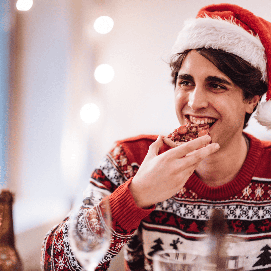 A smiling man wearing a festive Santa hat and holiday sweater enjoys a slice of pizza during a holiday gathering, highlighting the joy of mindful eating during the festive season.