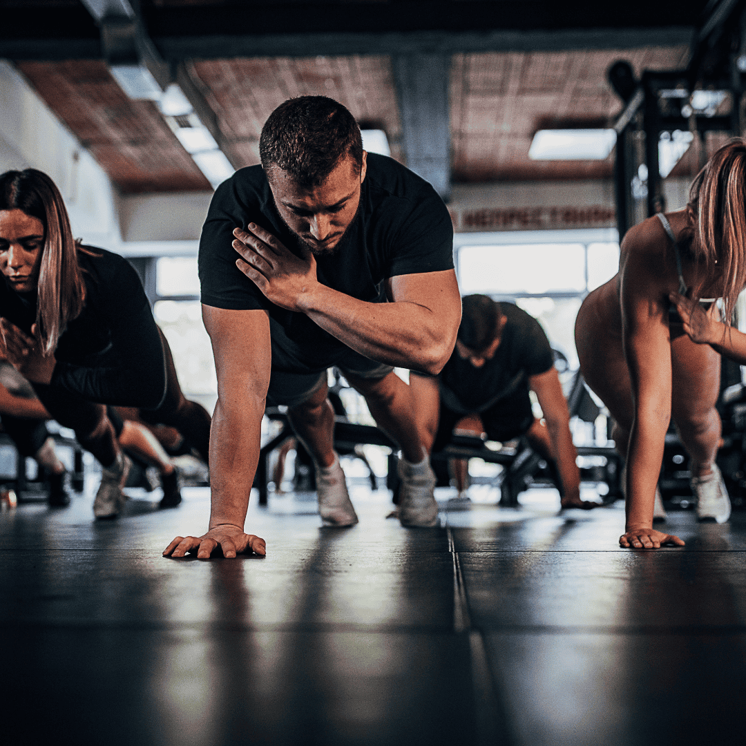 A group of individuals engaged in a high-intensity workout at a gym, with a man in the foreground performing a plank with a shoulder tap.