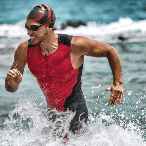 A male triathlete wearing a red and black wetsuit and swim cap emerges from the ocean, splashing through the water with a focused expression.